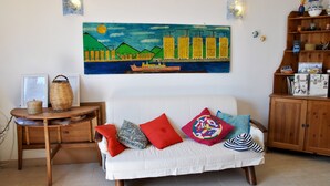 A living room with a white sofa, colorful cushions, a painting of a cityscape, and a wooden side table with a basket. - Casa Simon Nature and Sea (Sperlonga)