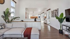 Un'ampia zona living con un divano, un tavolino e una pianta grande. In secondo piano, un angolo cottura con un tavolo da pranzo e sedie.