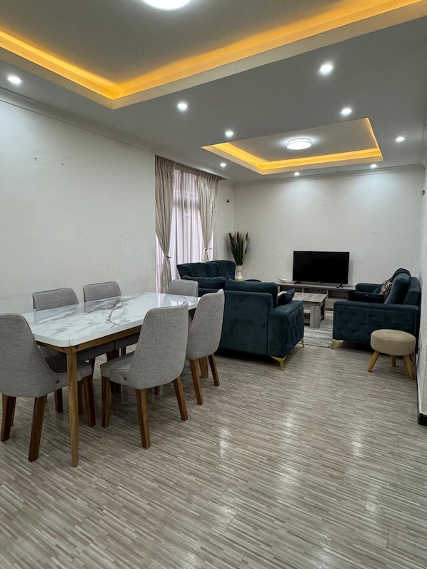 Un salon moderne avec un coin repas, comprenant une table en marbre, des chaises grises, des canapés foncés, un téléviseur à écran plat et un éclairage encastré au plafond.