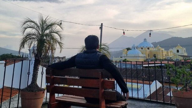 A person sitting on a bench overlooking a body of water with a mountain and buildings in the background. - Hotel Ciudad de los sueños (Antigua Guatemala)