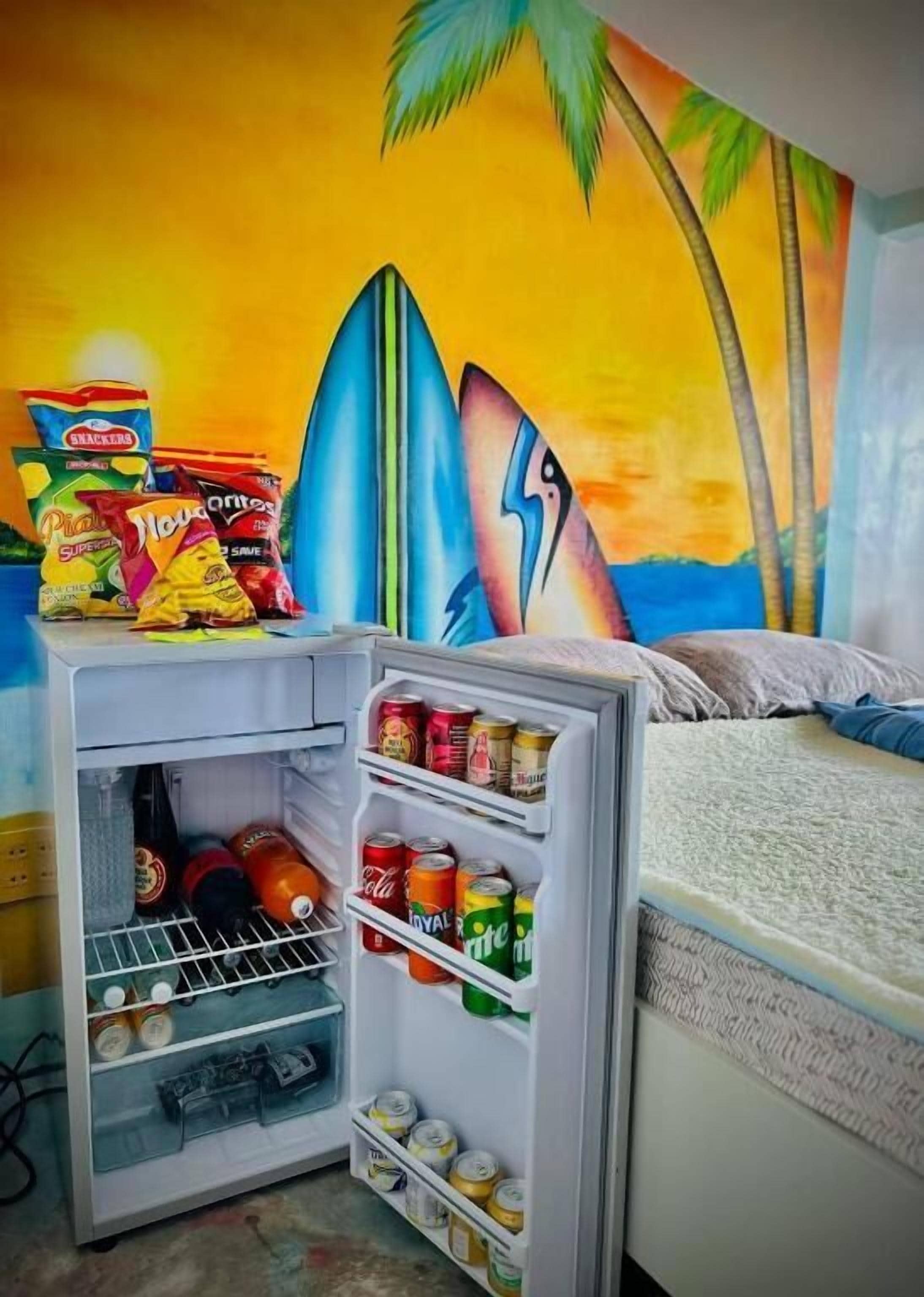 A compact refrigerator stocked with snacks and drinks, positioned in a room with a vibrant mural of a sunset and palm trees.