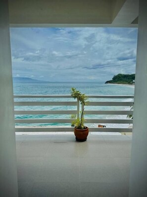 A balcony with a potted plant, overlooking a beach with clear blue waters and a distant mountain.
