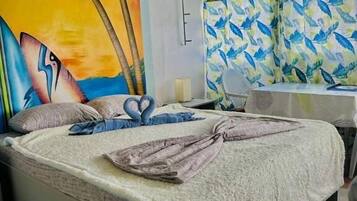 A hotel room with a bed, a mural of palm trees and a sunset, a shower curtain with a bird pattern, and a small table with a chair.