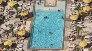 Luchtbeeld van een zwembad omringd door ligbedden en parasols, met mensen die zwemmen en ontspannen.