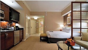 Hotelkamer met kitchenette, een bed met nachtkastje, zithoek met tafel en bloemen, en een groot raam met gebroken glaspanelen.