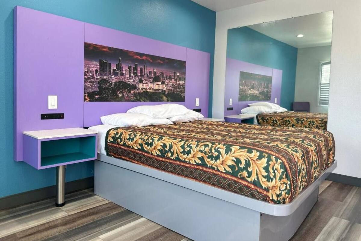 A modern bedroom with a cityscape mural, a large bed with a patterned bedspread, and a nightstand with a teal shelf.