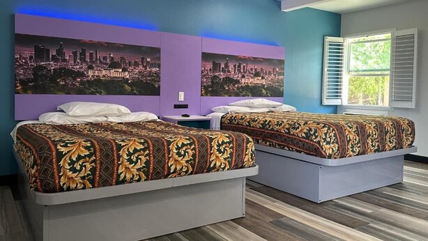 A hotel room with two beds, a cityscape mural on the wall, and a window with shutters.