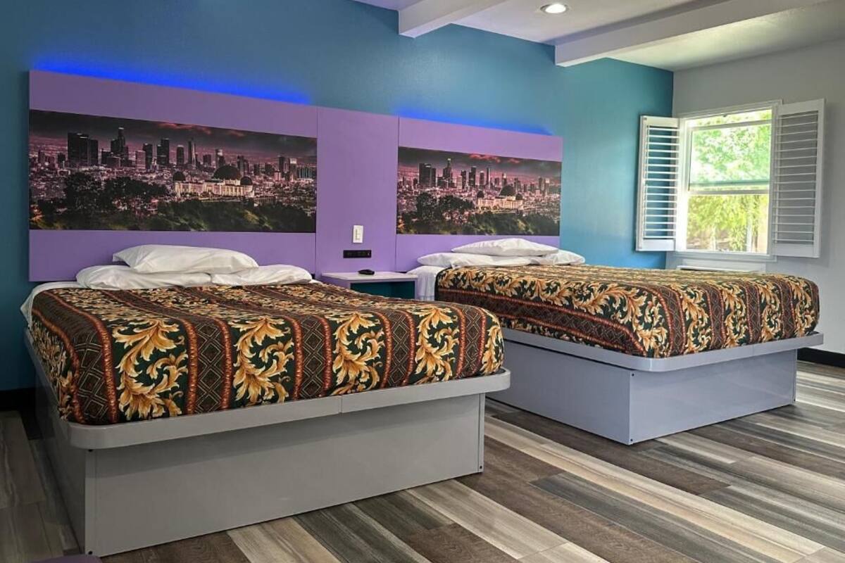 A hotel room with two beds, a cityscape mural on the wall, and a window with shutters.