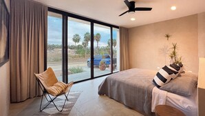 Une chambre avec un grand lit, une chaise, un ventilateur de plafond et une vue sur des palmiers et une voiture bleue à l’extérieur.