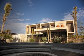 A modern building with a flat roof, large windows, and a courtyard with cacti and palm trees. - Kaánche House (Todos Santos)