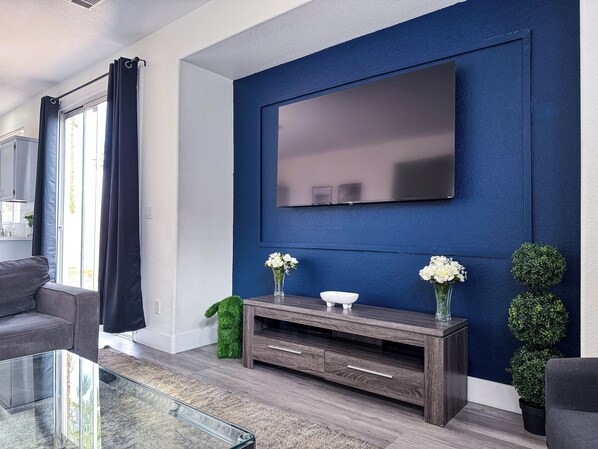 A modern living room with a large flat-screen TV mounted on a blue wall, a wooden entertainment unit with two vases of white flowers, and a glass coffee table.