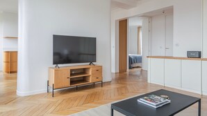 Un salon moderne avec un téléviseur à écran plat posé sur un meuble en bois, une table basse noire sur laquelle sont disposés des magazines, et un couloir menant à une chambre.