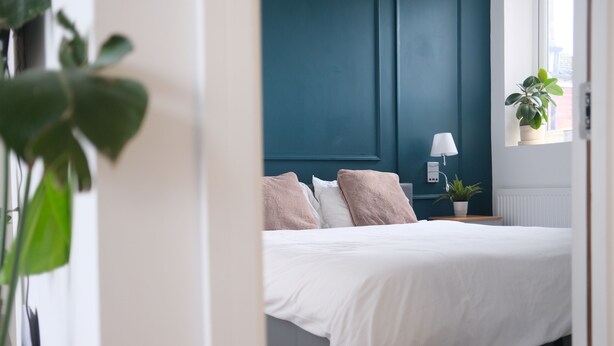 A bedroom with a bed, a nightstand, a lamp, and a window with a plant. - Brand New, Beautifully Designed Apartment in the Heart of Fulham (London)