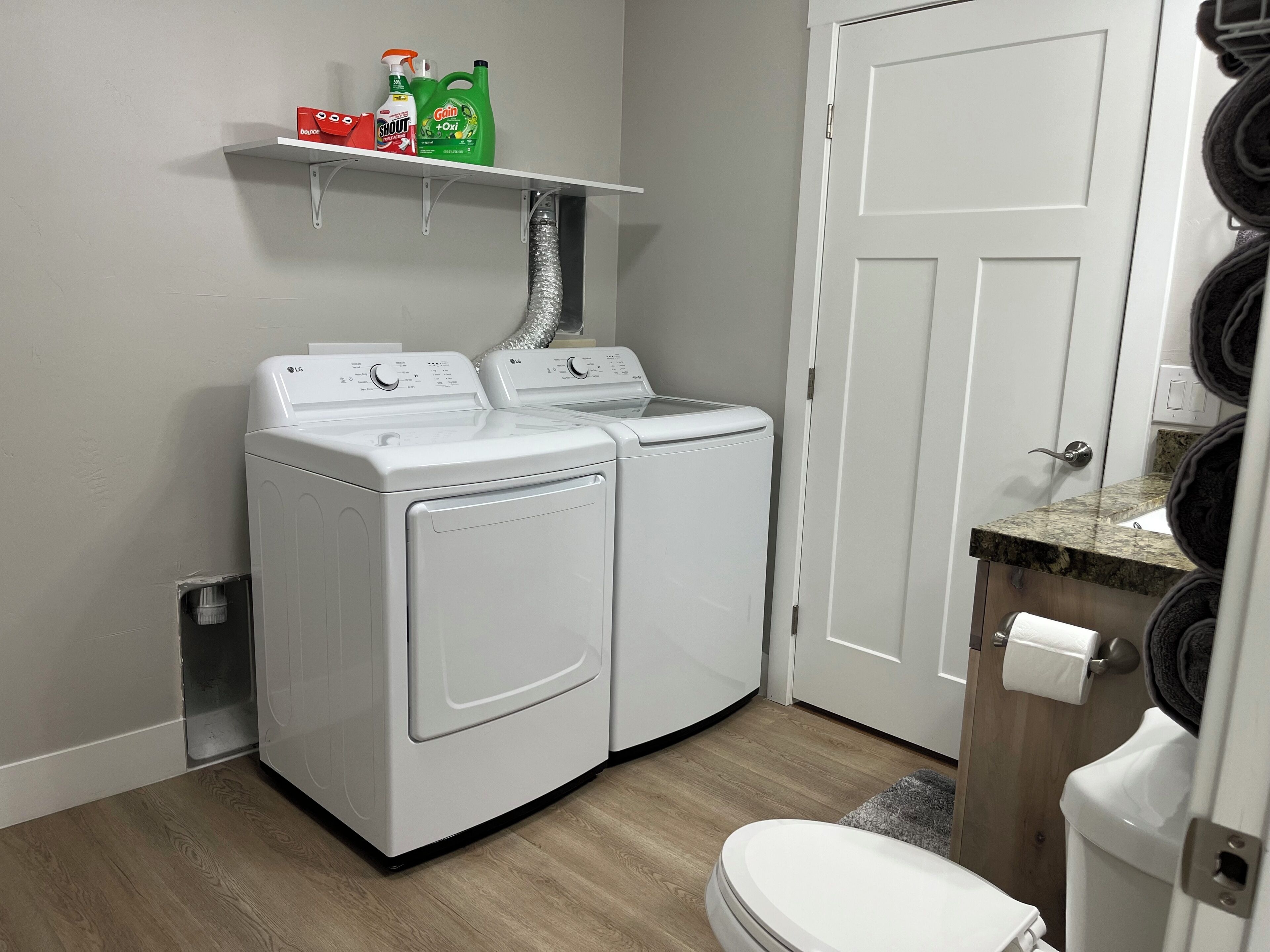 A laundry room with two white front-loading washing machines, a shelf with cleaning supplies, and a toilet with a towel dispenser.