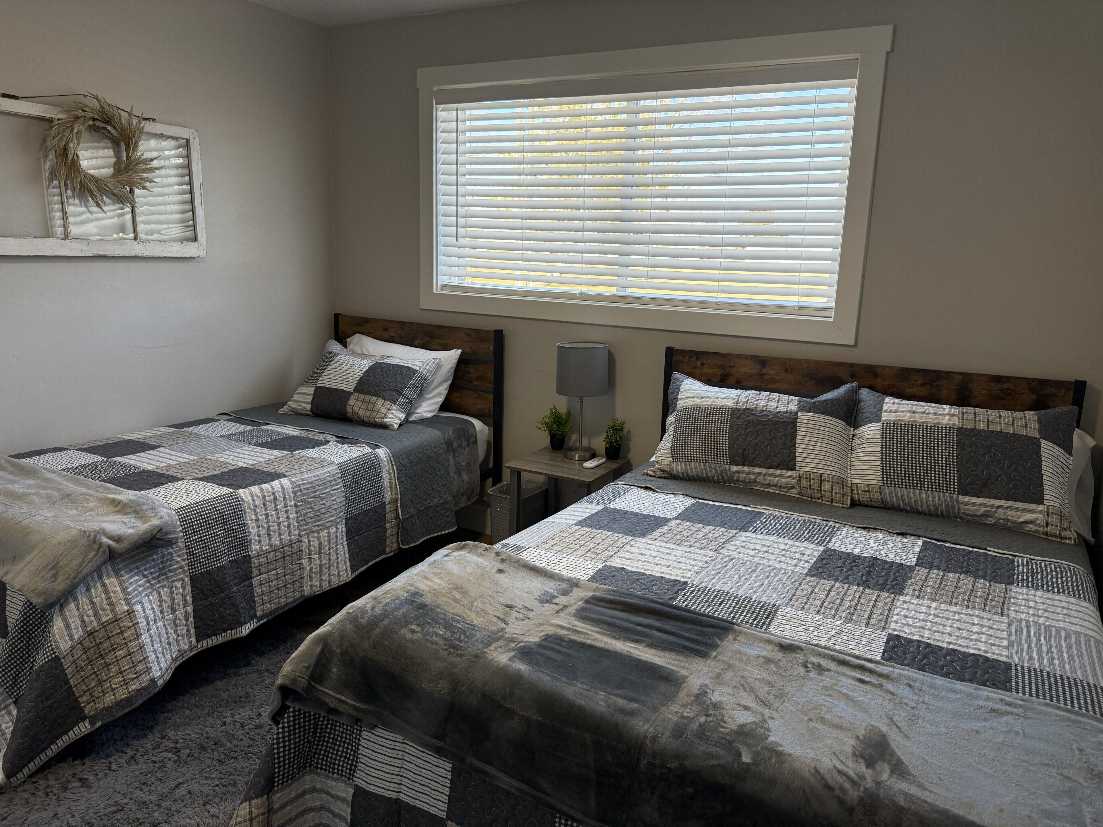 A bedroom with two beds, a window with blinds, a bedside table with a lamp, and a wall decoration.