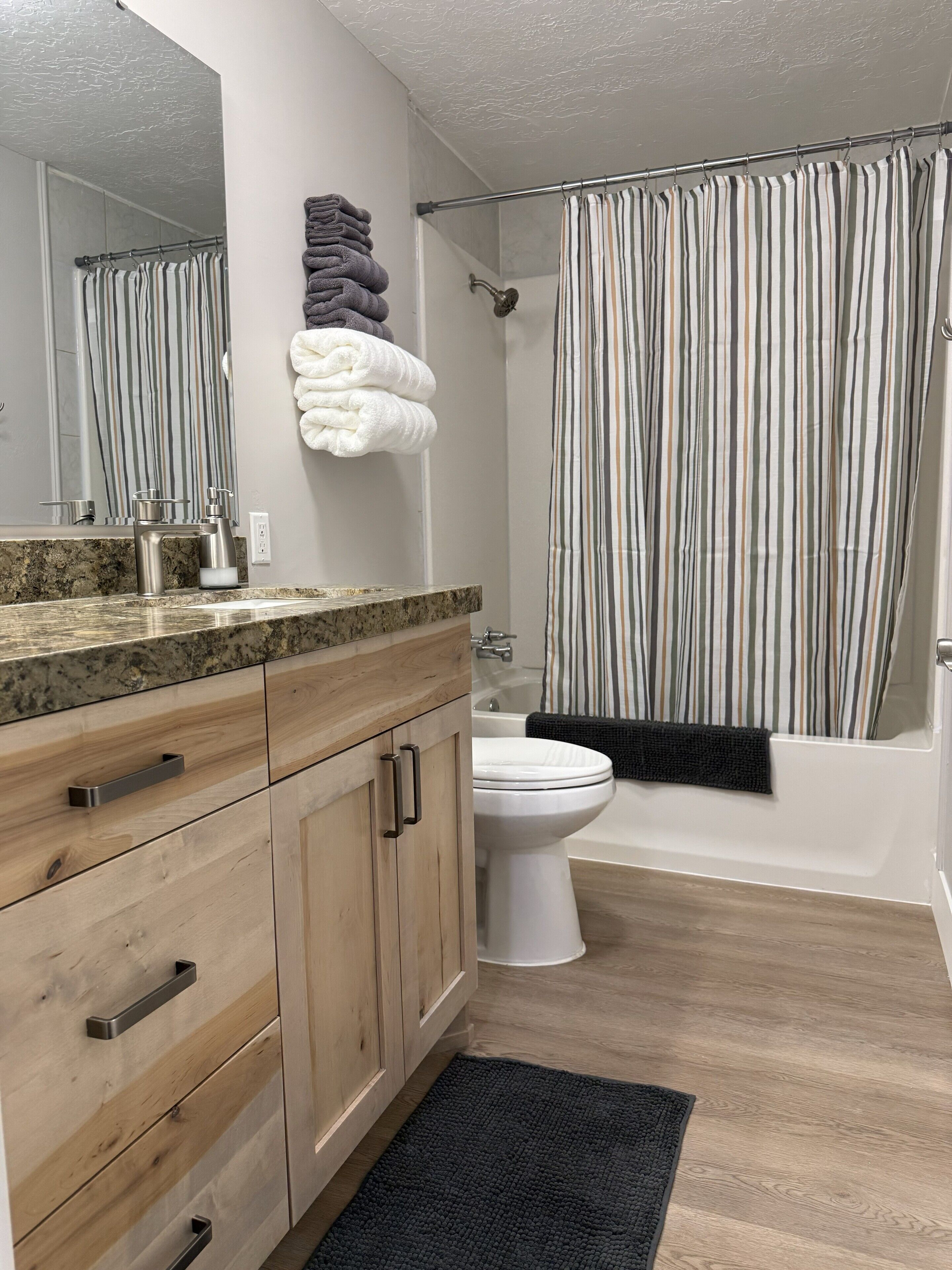 A modern bathroom with a wooden vanity, a white toilet, a bathtub, and a striped shower curtain.