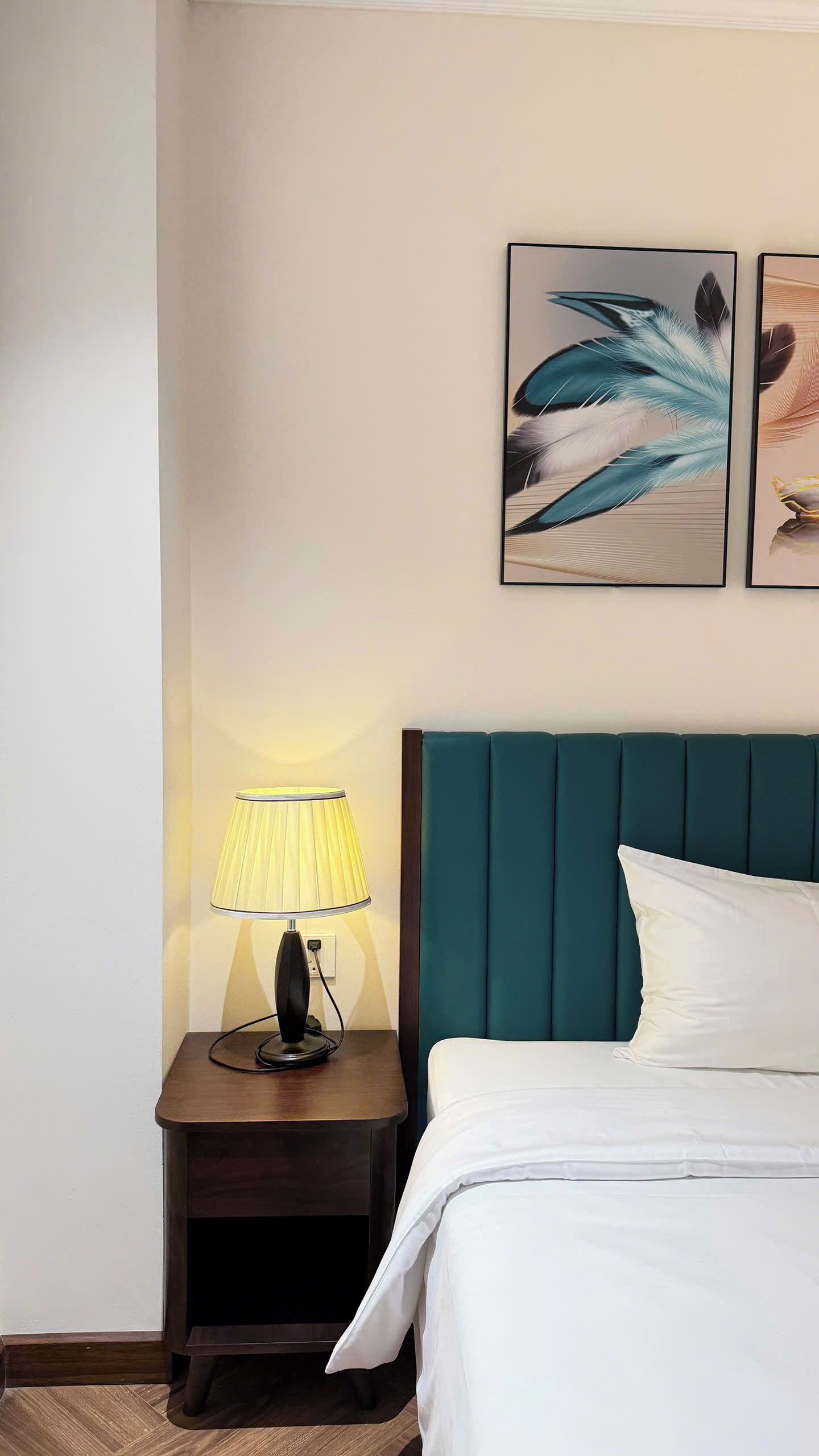 A hotel room with a bed, a nightstand with a lamp, and two framed artworks on the wall.