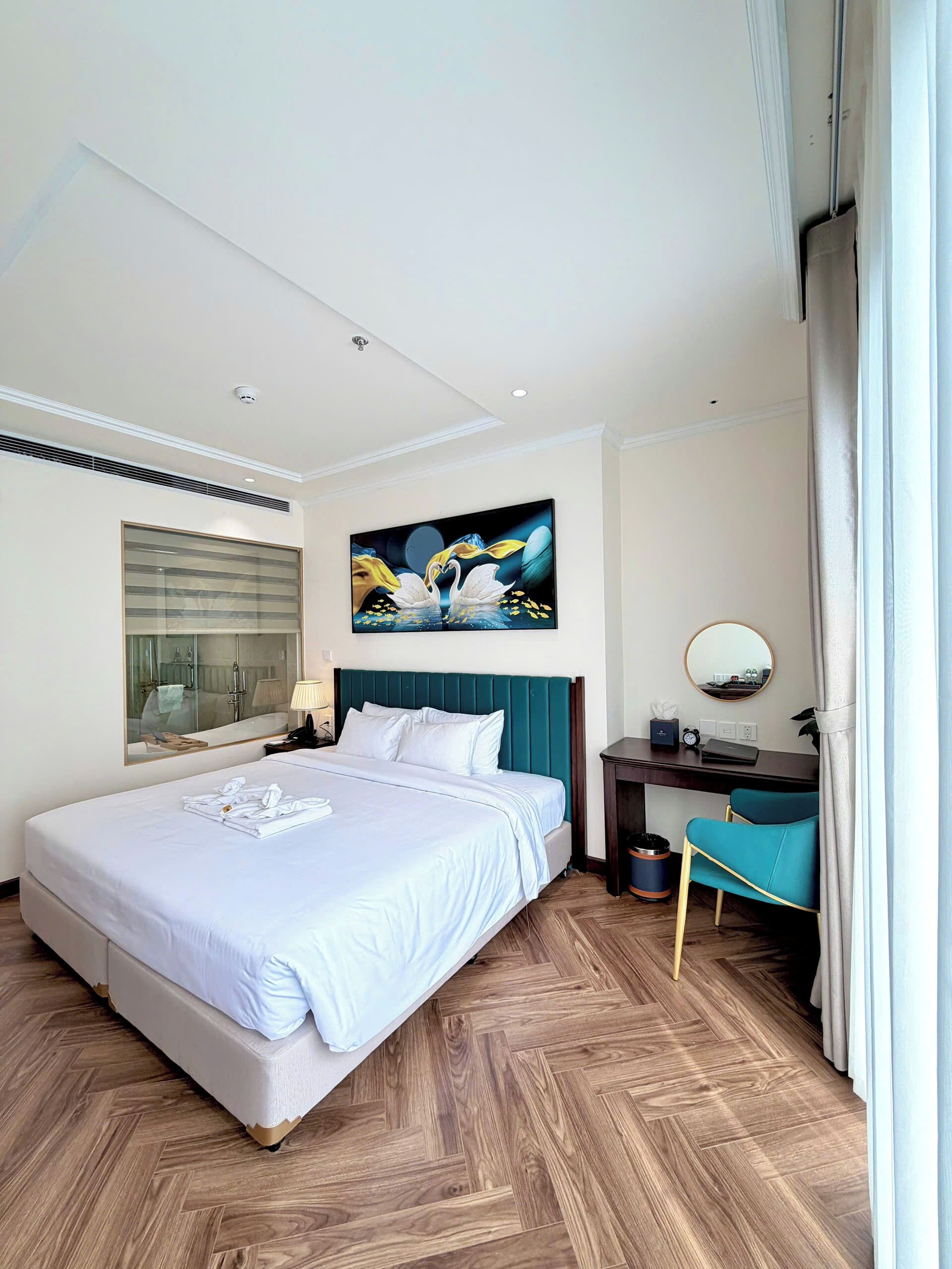 A modern hotel room with a large bed, a desk, a chair, a mirror, and a painting on the wall.