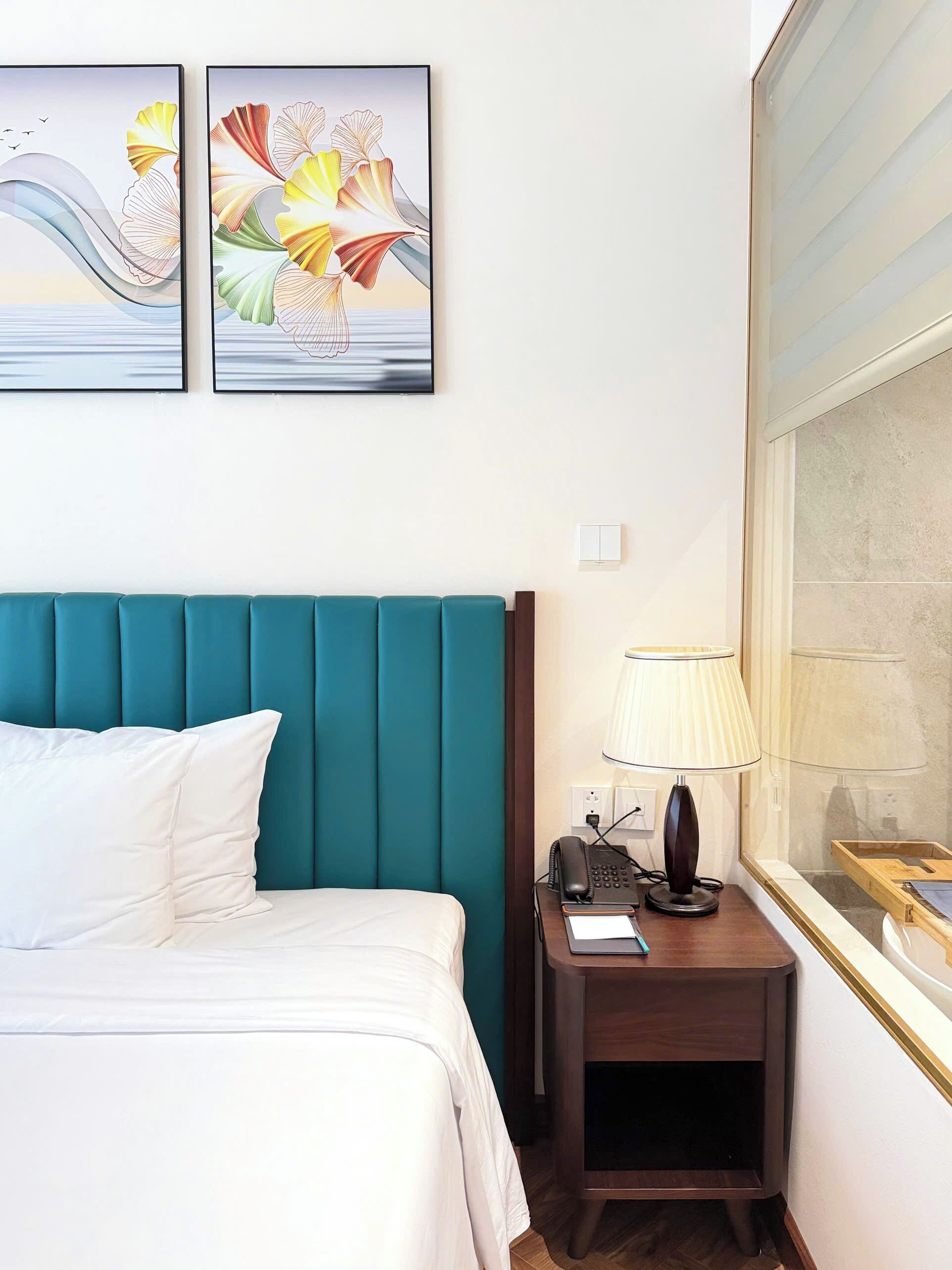 A hotel room with a teal upholstered headboard, a white bed with pillows, a bedside table with a lamp, and two framed artworks above the bed.