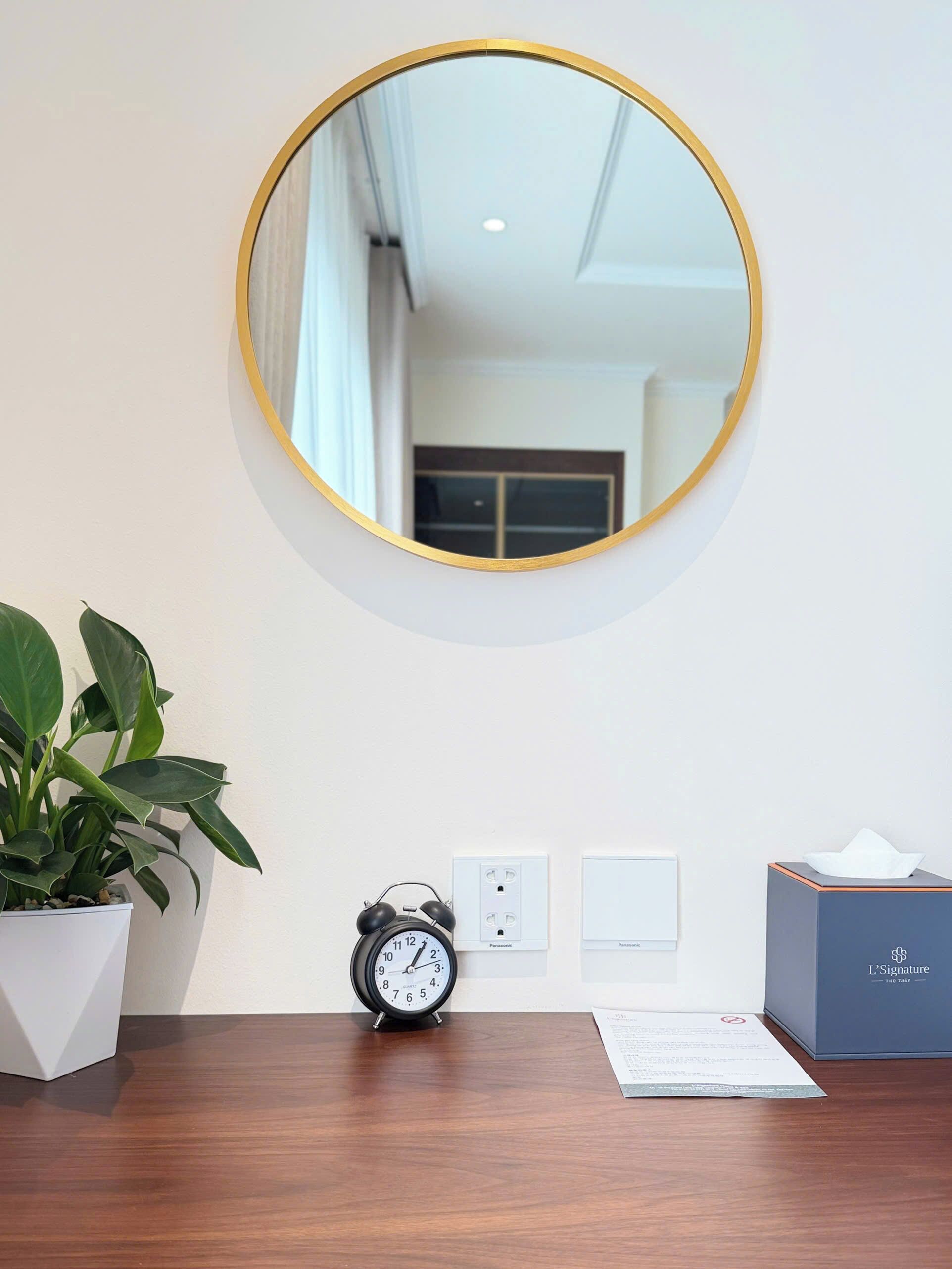 A round mirror with a gold frame, a black alarm clock, a white power outlet, a tissue box, and a potted plant on a wooden desk.
