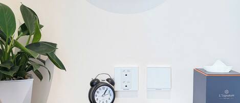 A round mirror with a gold frame, a black alarm clock, a white power outlet, a tissue box, and a potted plant on a wooden desk.