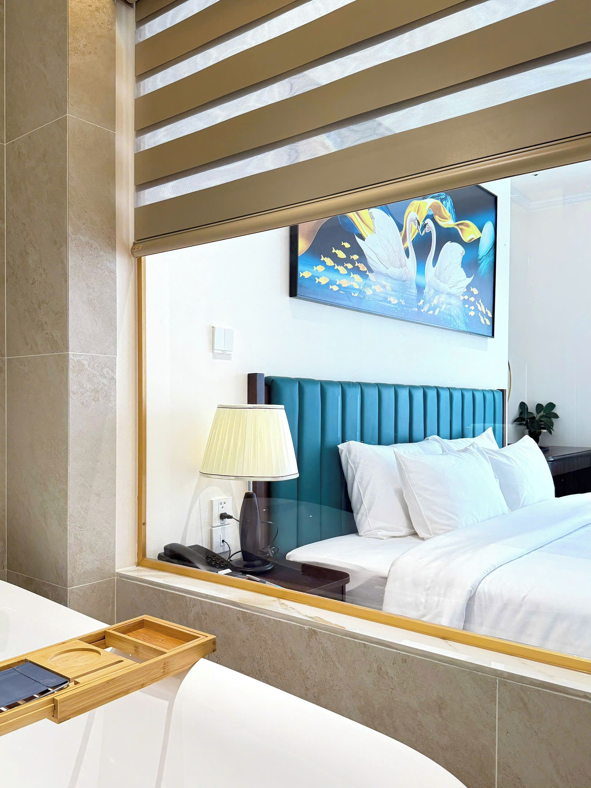 A modern hotel room with a large bed, a bedside table with a lamp, a window with a view of the sea, and a painting on the wall.