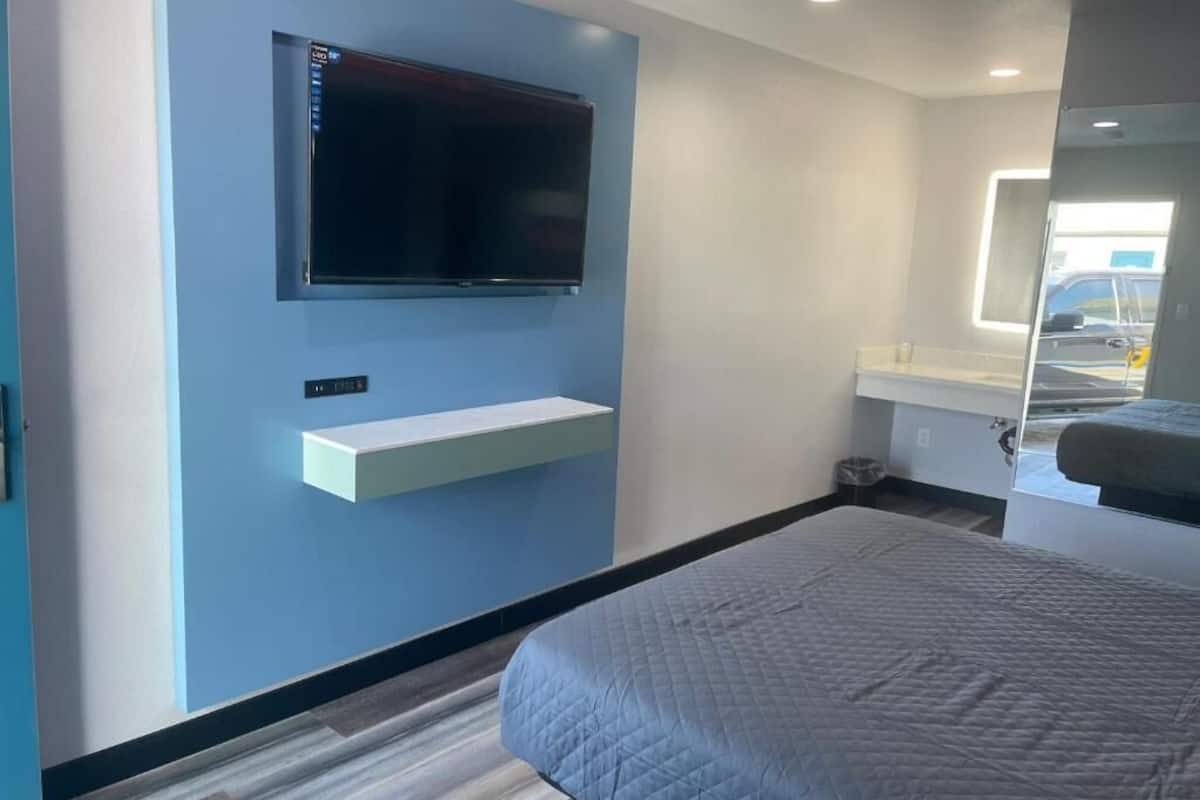 Un dormitorio con cama, televisor montado, una pequeña estantería y ventana con vistas al exterior.