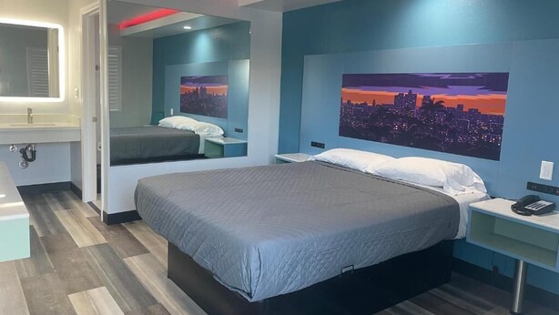 A modern hotel room with a large bed, a vanity with a mirror, a nightstand, and a cityscape painting on the wall. - La Palma Motel - South Gate - LA area (South Gate)