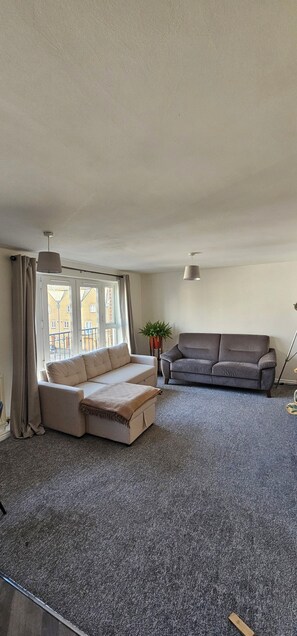 Un salon avec un canapé d’angle beige et un canapé gris, un sol recouvert d’une moquette, de grandes fenêtres avec des rideaux et une plante en pot.
