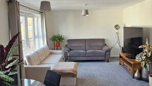 A modern living room with a grey sofa, a glass dining table with chairs, a TV, and a floor lamp.