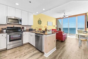 A modern kitchen with white cabinets, stainless steel appliances, and a wooden floor. There is a dining area with a red chair and a table. - Voted Top Beachfront Condo! Views, Hot Tubs, Pools (Gulf Shores)