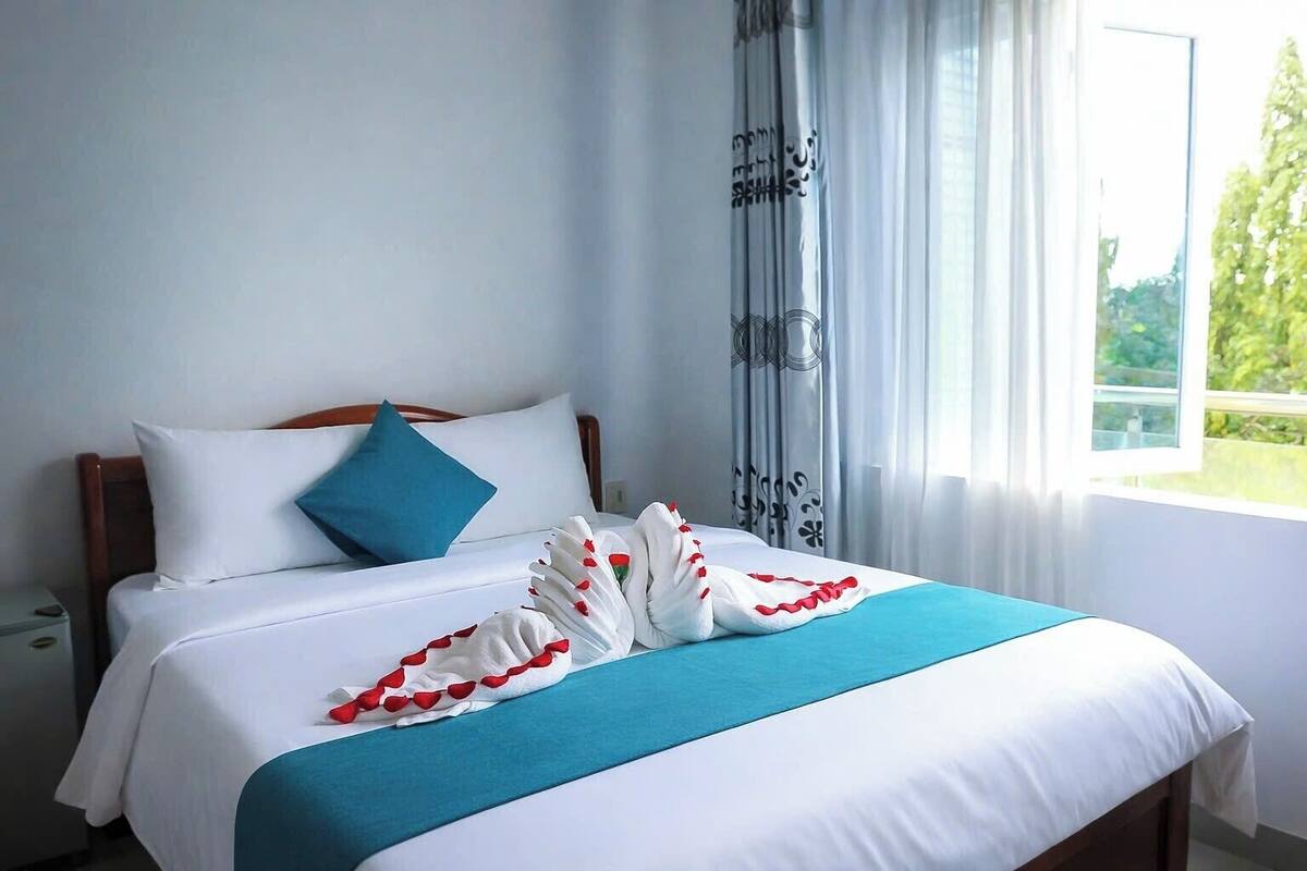 Une chambre d’hôtel avec un lit, un oreiller bleu, une couverture blanche et une fenêtre avec des rideaux.