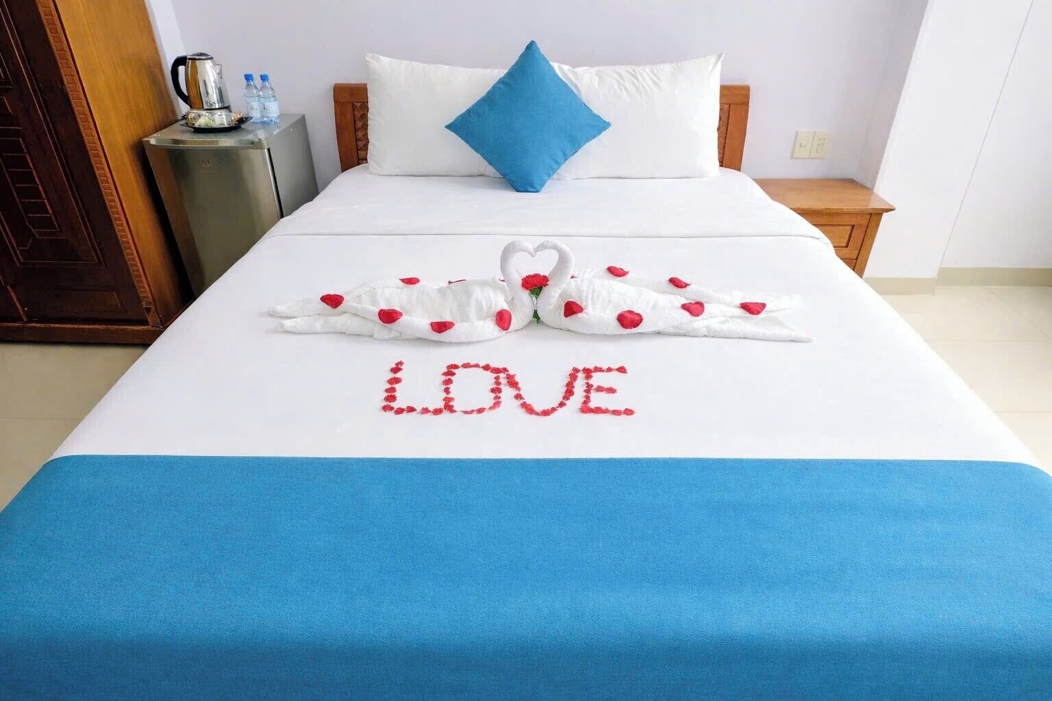 A hotel room with a bed, a blue bedspread, a decorative swan and heart arrangement spelling "LOVE" on the bed, a blue pillow, and a small bedside table with a kettle and water bottles.