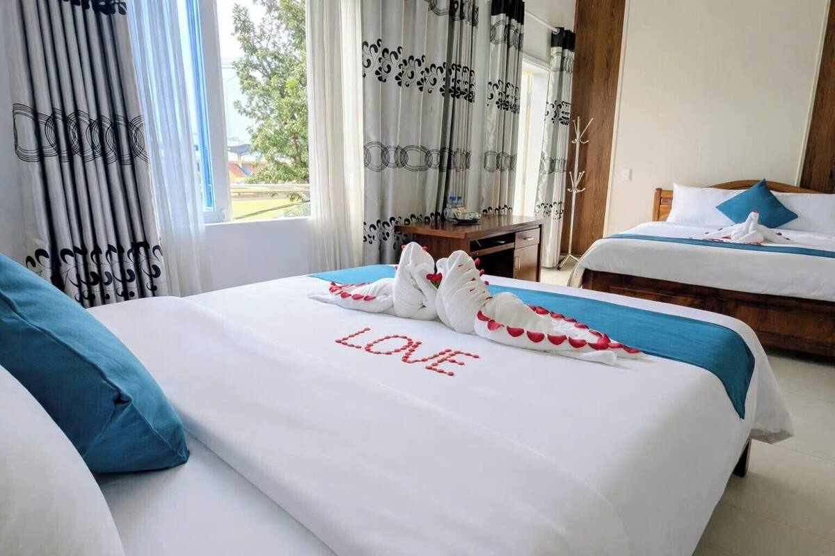 Une chambre d’hôtel avec deux lits, dont l’un est décoré de motifs de cygnes et sur lequel est inscrit le mot « AMOUR » sur la couvre-lit, et qui offre une vue sur les arbres à l’extérieur.