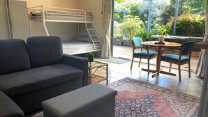 A compact room with a bunk bed, a sofa, a small table with chairs, and a view of a garden through large windows. - Cozy 1 bedroom unit nestled in a lush sub tropical garden located in Kerikeri. (Kerikeri)