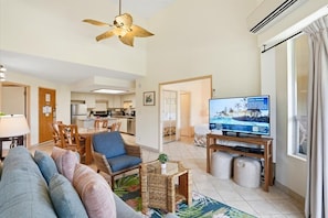 A living room with a sofa, a television, a ceiling fan, and a kitchen in the background. - Maui Banyan condo w/ BBQ grill and pool (Kihei)