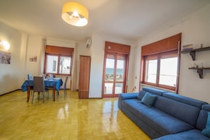 Une pièce avec un canapé bleu, une table à manger avec des chaises, un luminaire au plafond et une vue sur l’extérieur, visible à travers une porte.