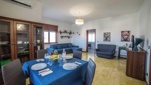 A dining area with a blue tablecloth, a dining table set with plates, glasses, and a bottle. There are two sofas, a cabinet with glass doors, and a ceiling fan. - Tramonto D'Oro Apartment Near The Beach, Porto Cesareo, Italy (Porto Cesareo)