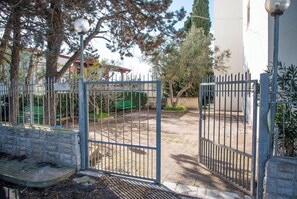 Ein verschlossener Eingangsbereich mit einem Metallgitter, einem gepflasterten Weg und einer Bank.