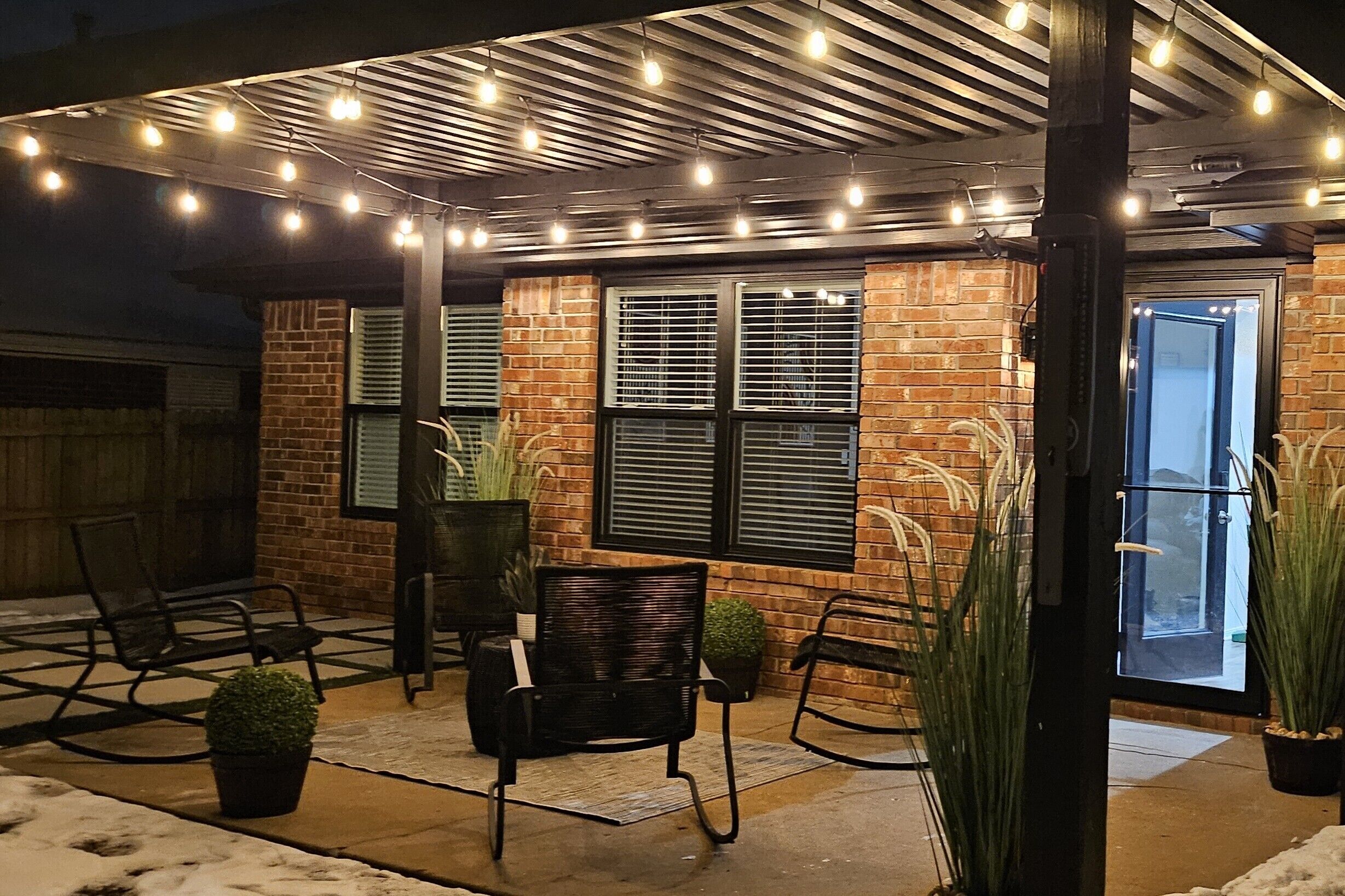 An outdoor patio area with brick walls, black metal chairs, potted plants, and string lights.