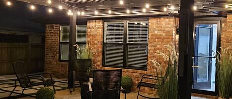 An outdoor patio area with brick walls, black metal chairs, potted plants, and string lights.