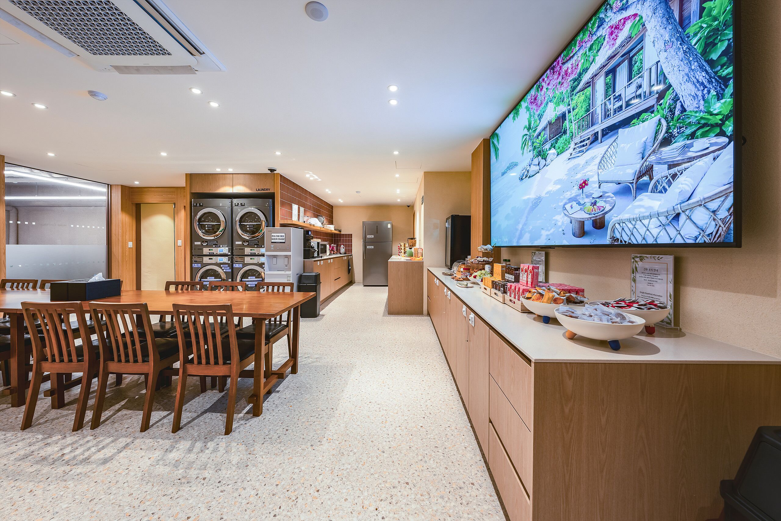 A hotel room with a dining area, a kitchen, a laundry room, and a large TV screen displaying a tropical scene.