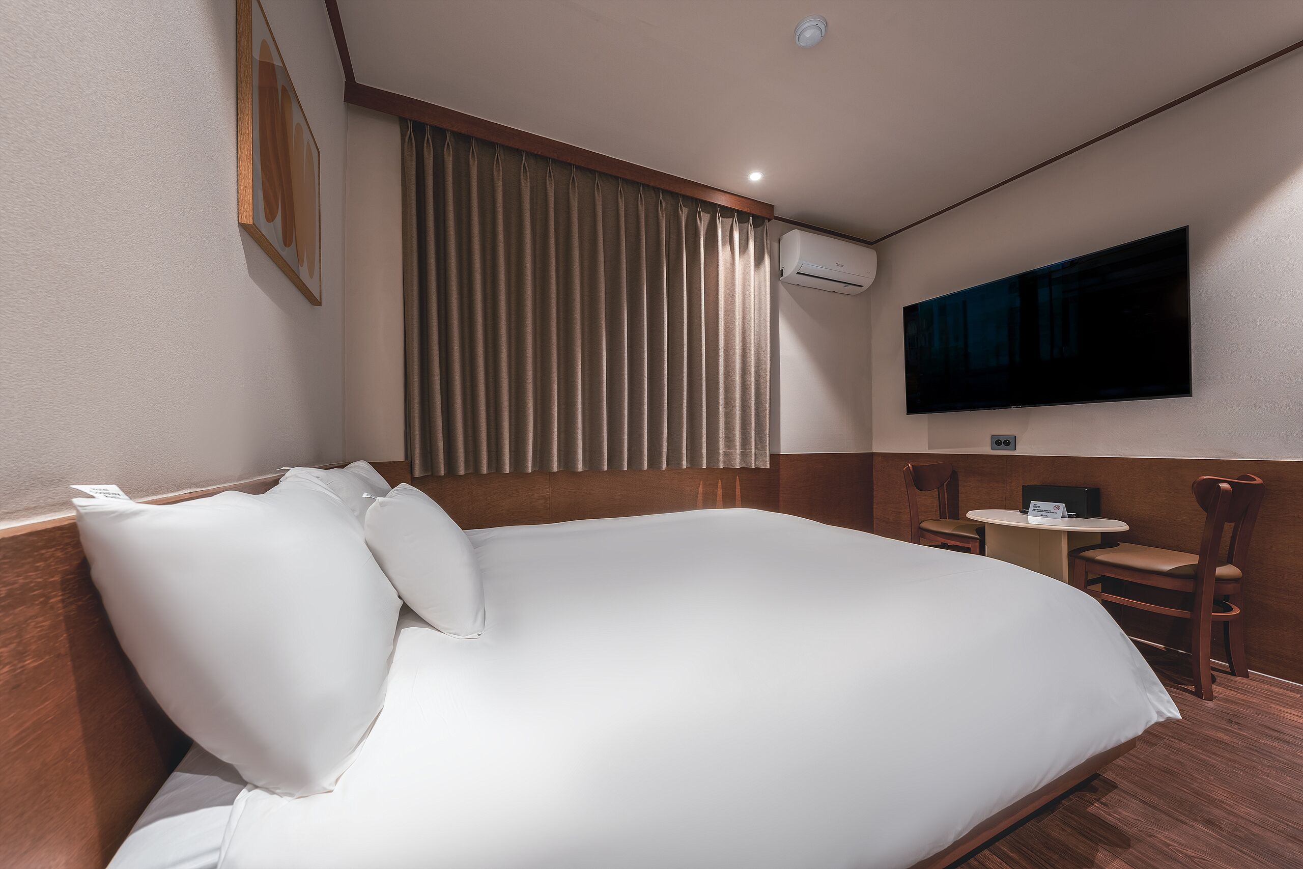 Habitación de hotel con una cama grande, un televisor de pantalla plana en la pared, un equipo de aire acondicionado y una pequeña mesa con una silla.