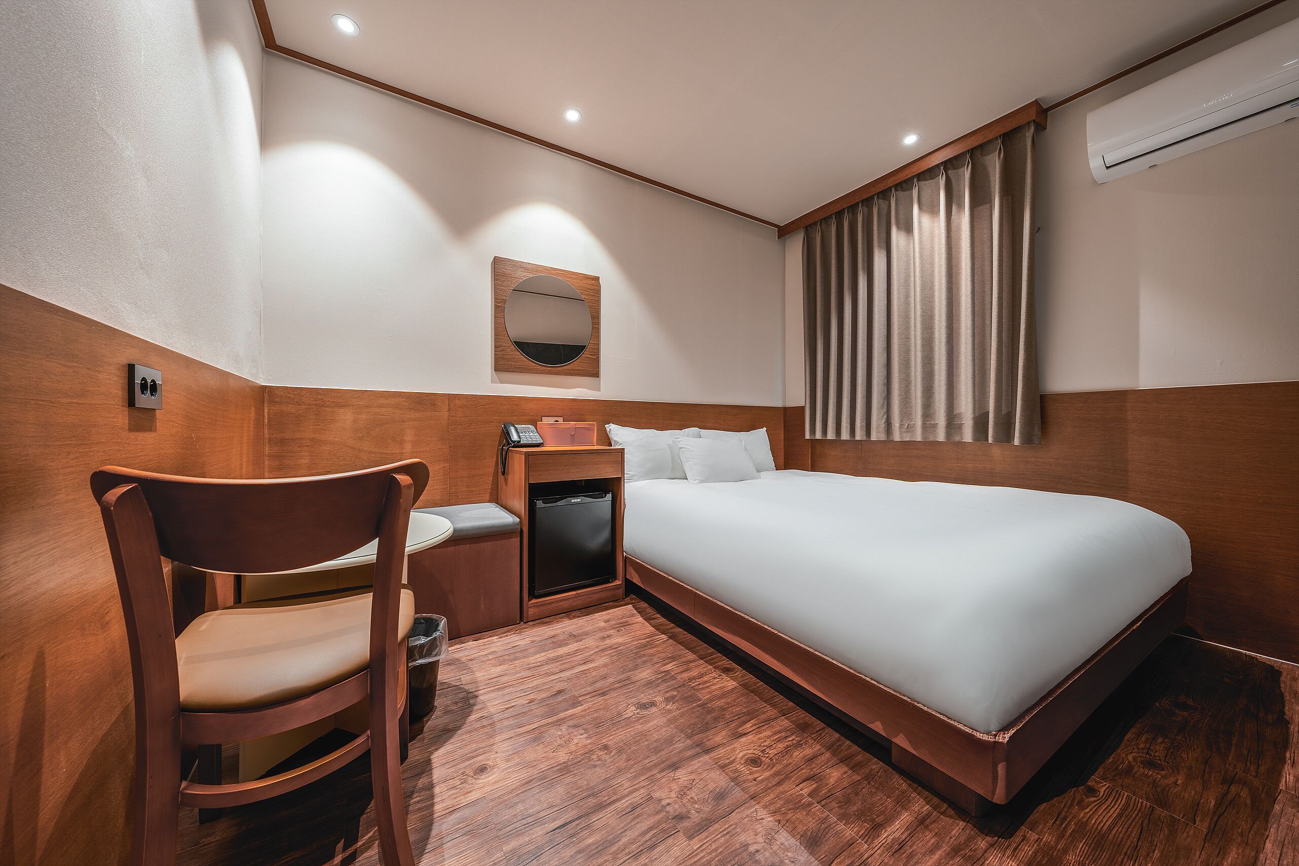 A hotel room with a bed, a chair, a small table with a phone, a mirror, and a window with curtains.