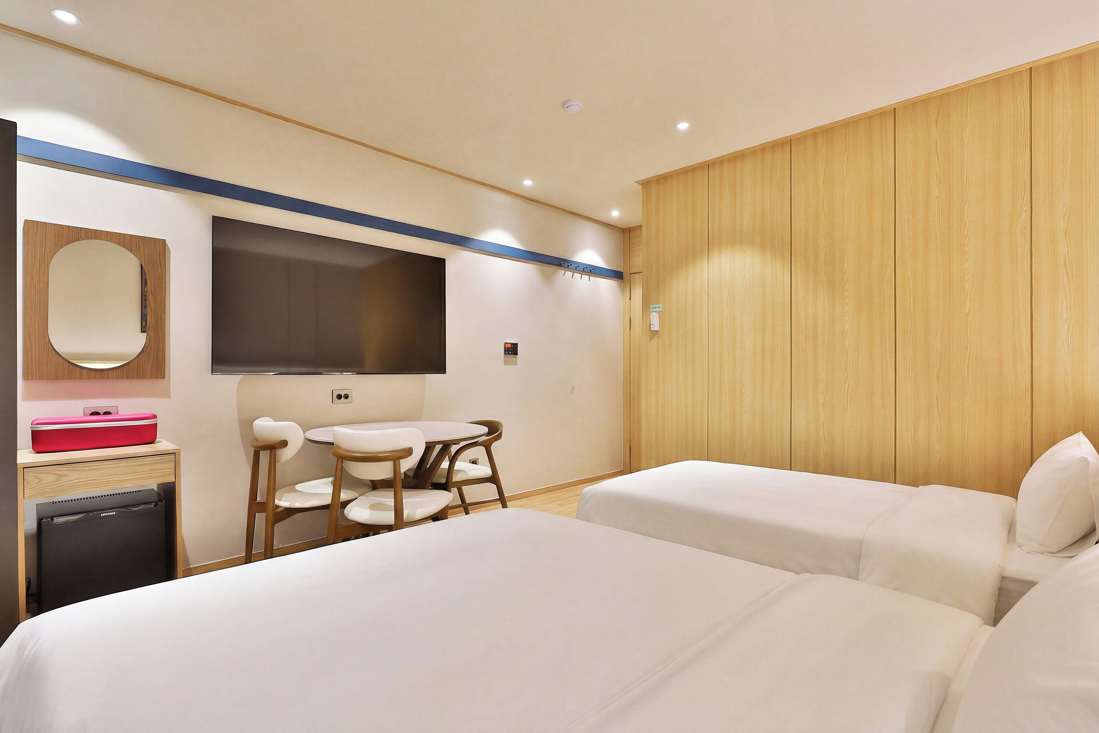 A hotel room with two beds, a desk with a chair, a small table, a mirror, and a TV.