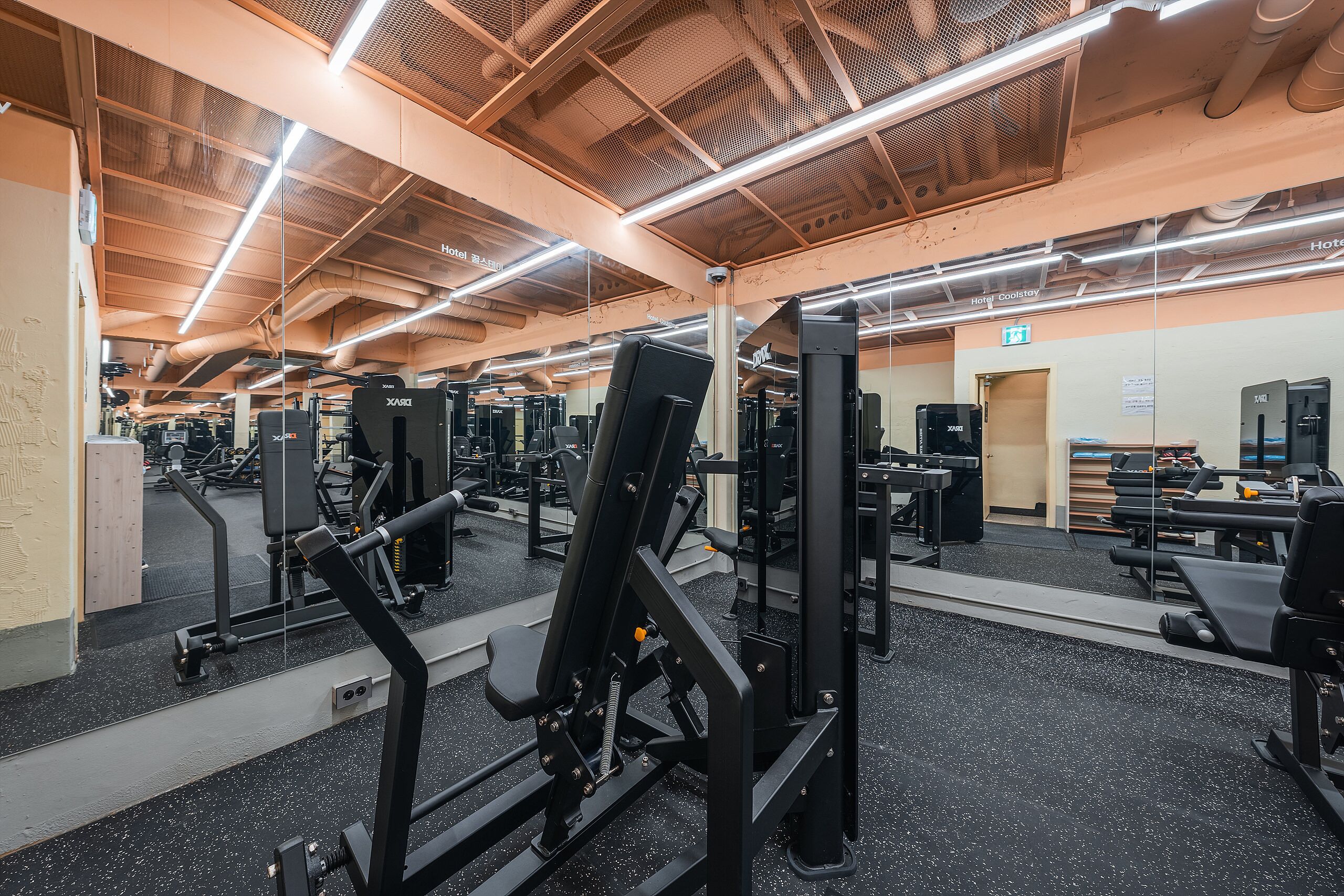 Un gimnasio con equipos de ejercicio variados, un espejo grande y un techo con conductos a la vista.