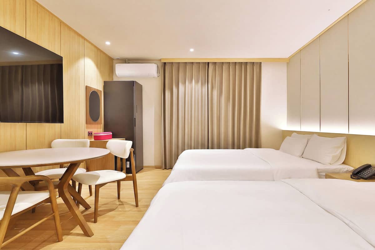 A hotel room with a wooden floor, a round dining table with chairs, two beds, a wardrobe, a TV, and a curtain.