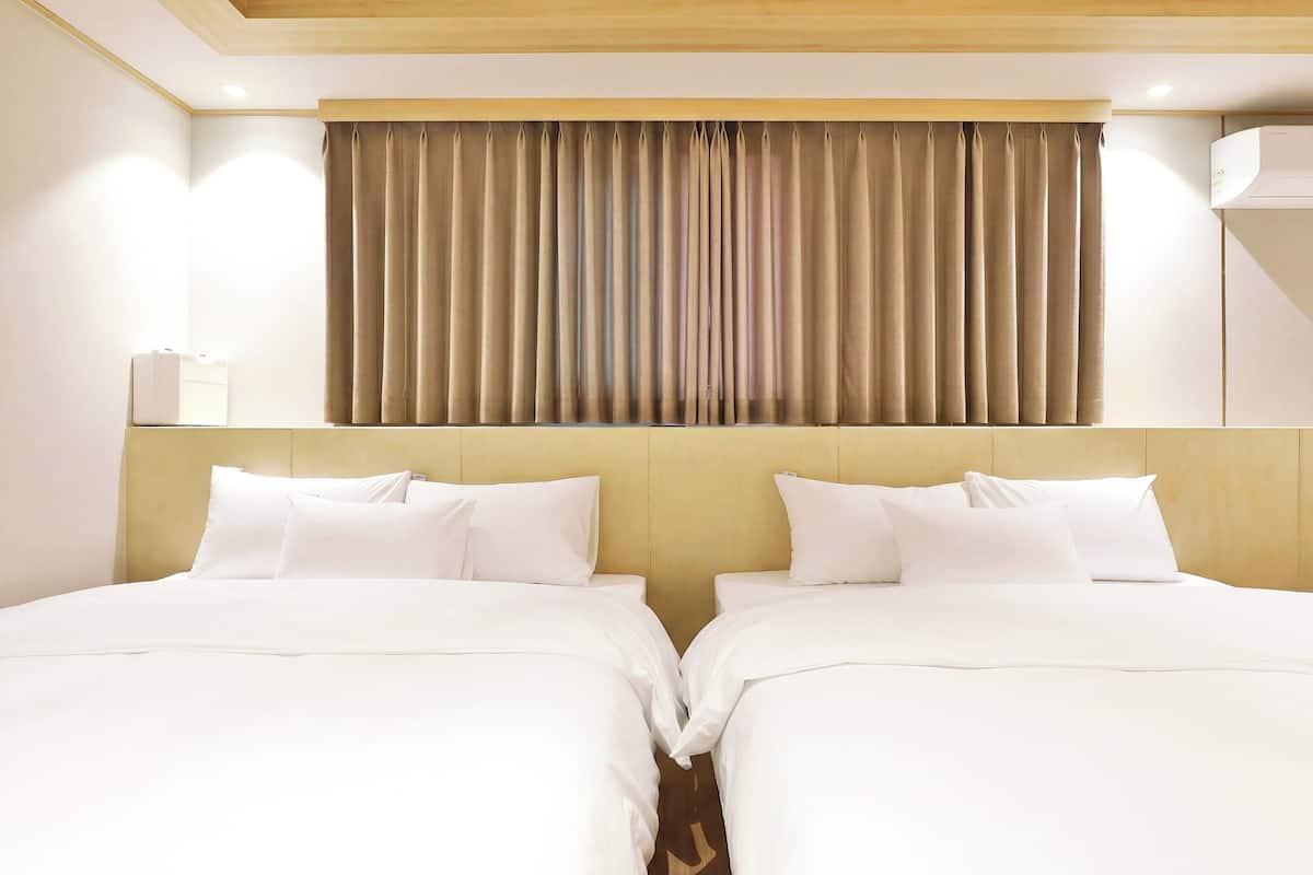 A hotel room with two beds, beige curtains, a wooden headboard, and a white wall-mounted air conditioner.