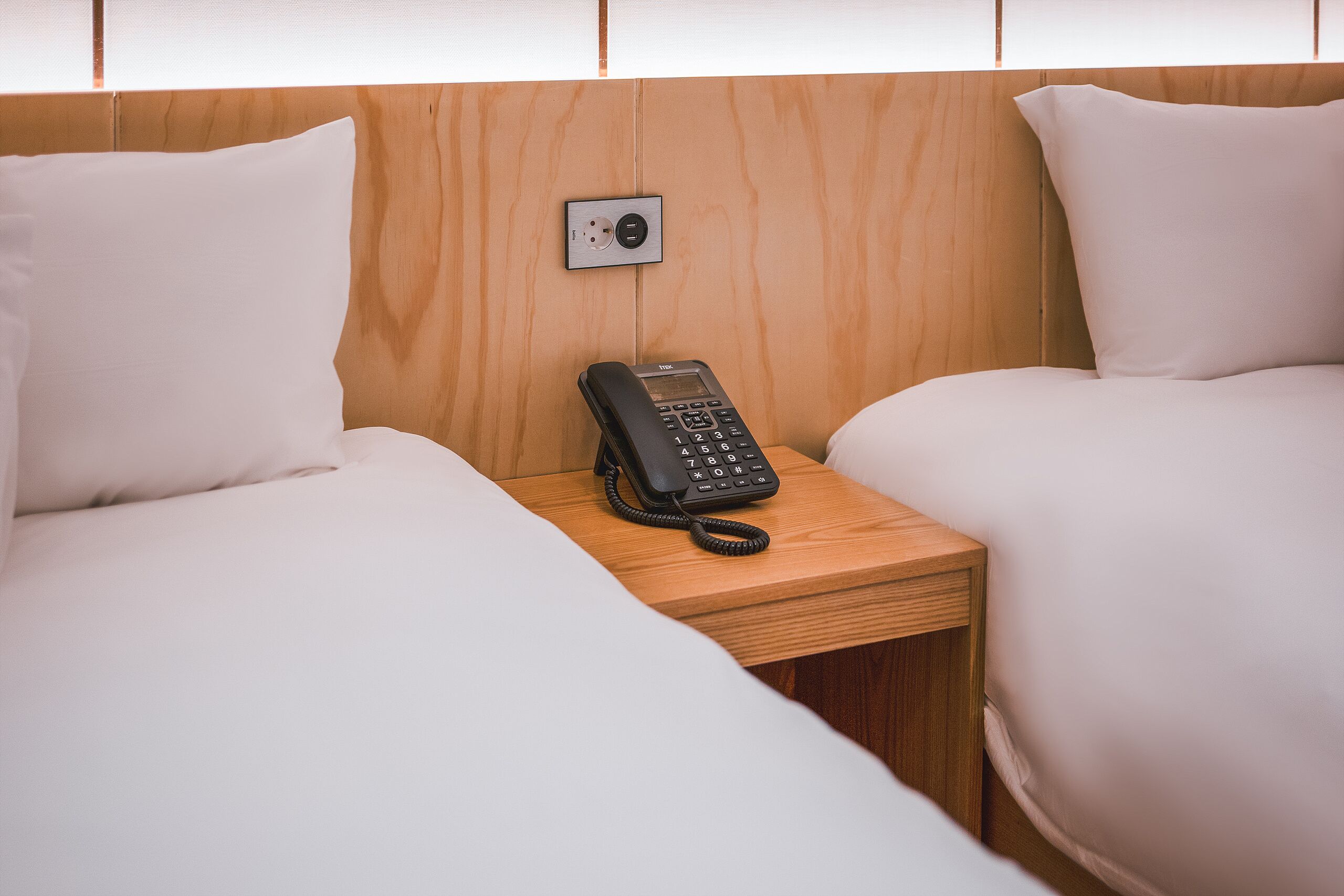 Habitación de hotel con dos camas, cabecera de madera, mesita de noche con teléfono y enchufe.