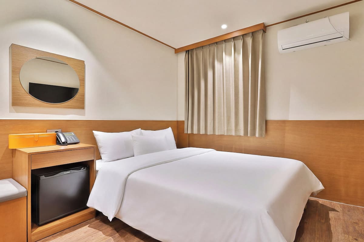 A hotel room with a bed, a bedside table with a phone and a small appliance, a mirror, and an air conditioning unit.